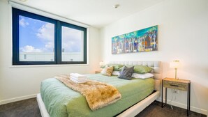A bedroom with a large bed, a window, a painting, and a nightstand with a lamp.