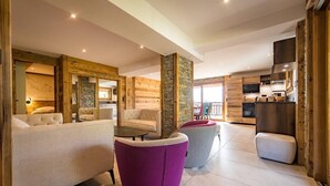 Un intérieur moderne avec un espace de vie comprenant un canapé, deux fauteuils et une table basse. On aperçoit à l’arrière-plan un coin cuisine avec des meubles en bois et un téléviseur.