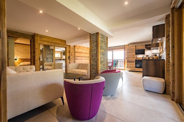 Un interior moderno con una sala de estar que incluye un sofá, dos sillones y una mesa de centro. Se observa en el fondo una zona de cocina con armarios de madera y un televisor.