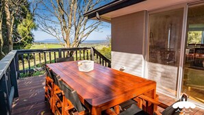 Une terrasse en bois avec une table et des chaises, une porte vitrée donnant sur un espace intérieur, et une vue sur un paysage arboré avec un horizon lointain.