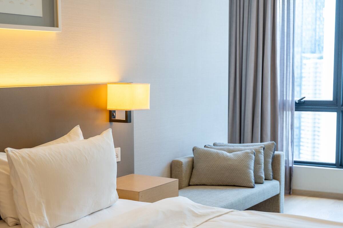 Quarto de hotel com cama, mesa de cabeceira, luminária e janela com cortinas.