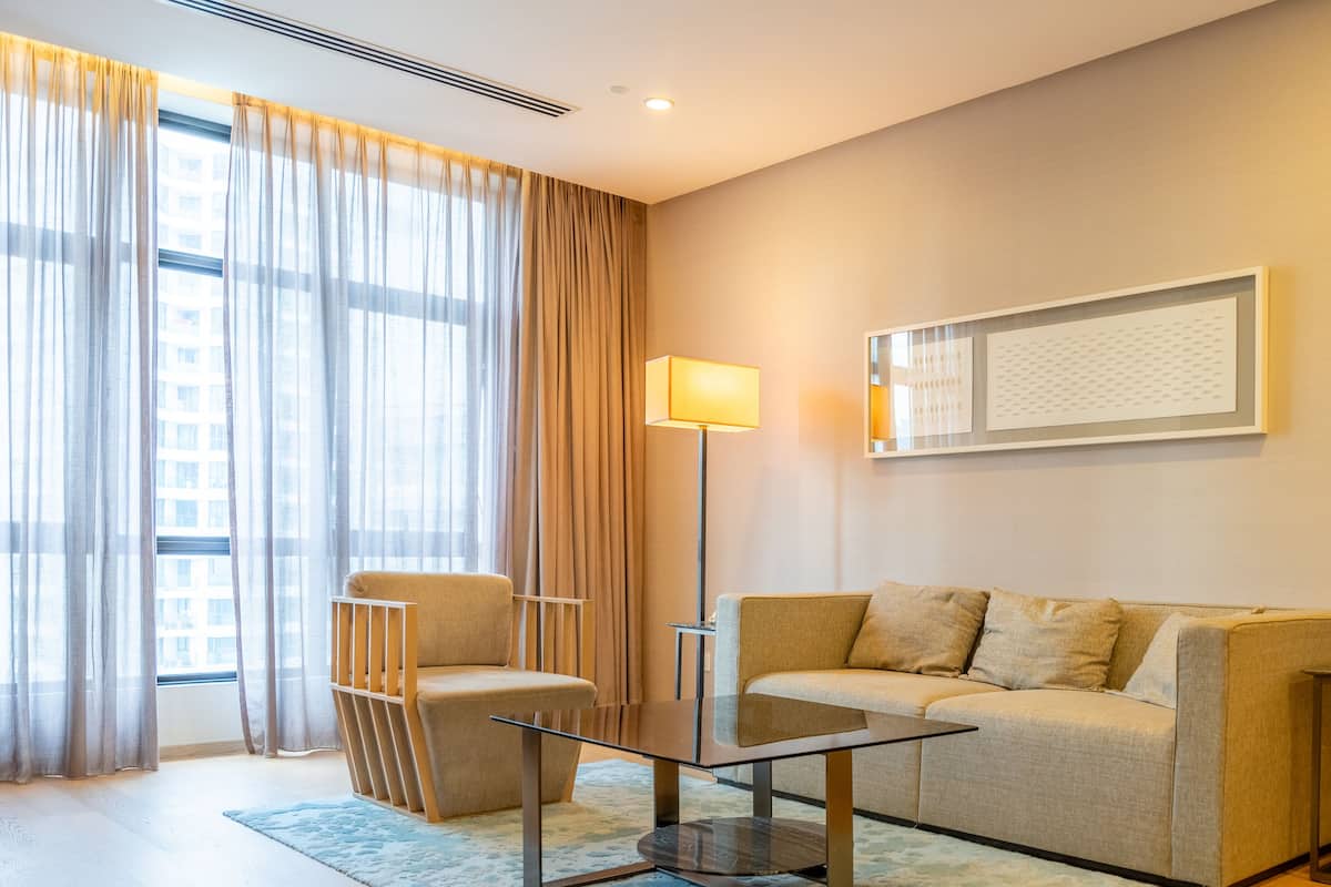 Quarto de hotel moderno com sofá, mesa de centro de vidro, luminária de chão e uma janela grande com cortinas.