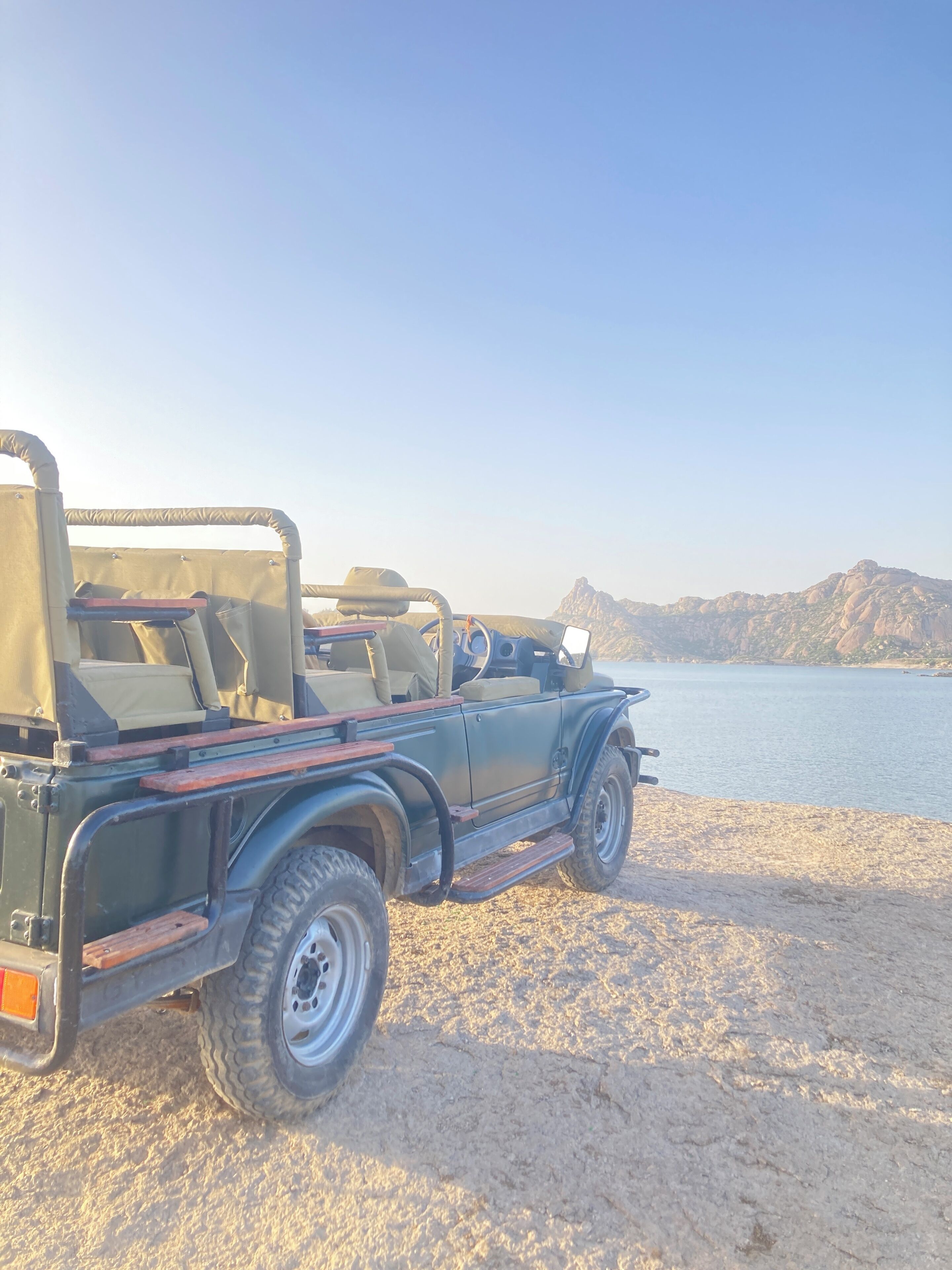 Een cabrio Jeep staat geparkeerd op een zandstrand, met een helder uitzicht op het water en de bergen op de achtergrond.