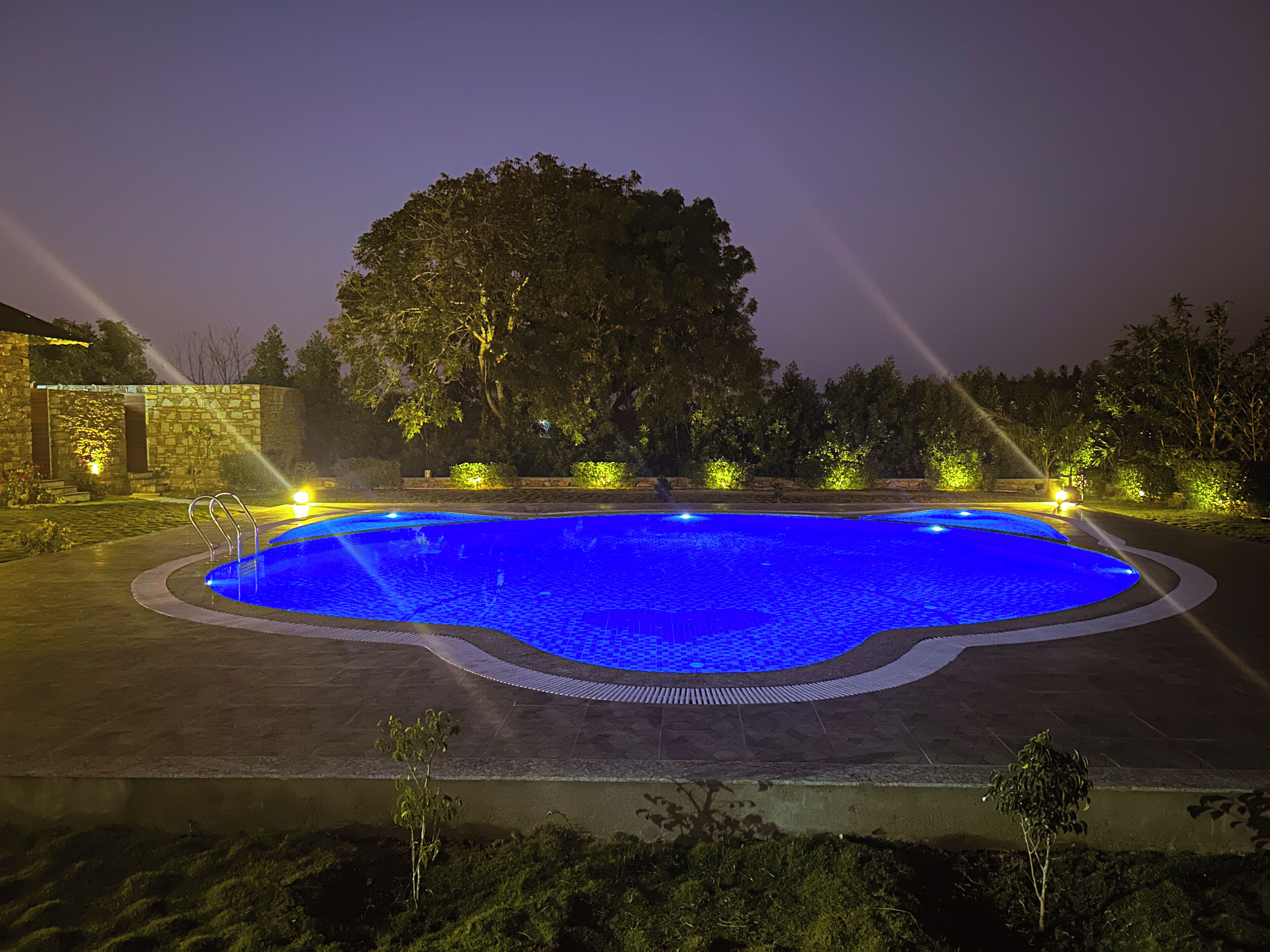 Une piscine nocturne aux reflets bleutés, entourée d’une terrasse dallée et éclairée par des projecteurs.