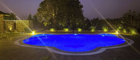 Une piscine nocturne aux reflets bleutés, entourée d’une terrasse dallée et éclairée par des projecteurs.