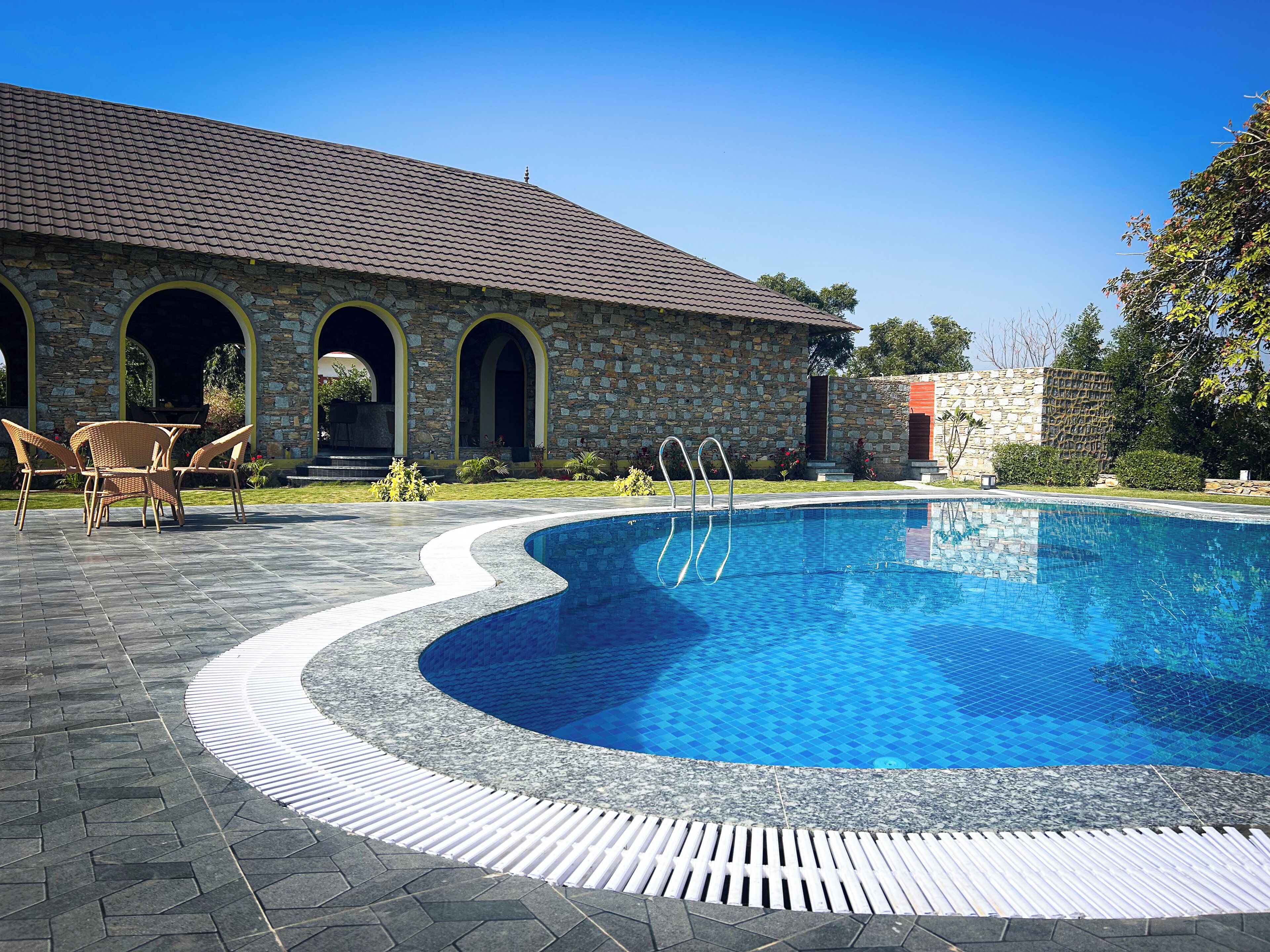 Una piscina con un edificio de piedra al fondo, un patio con sillas y un cielo azul despejado.
