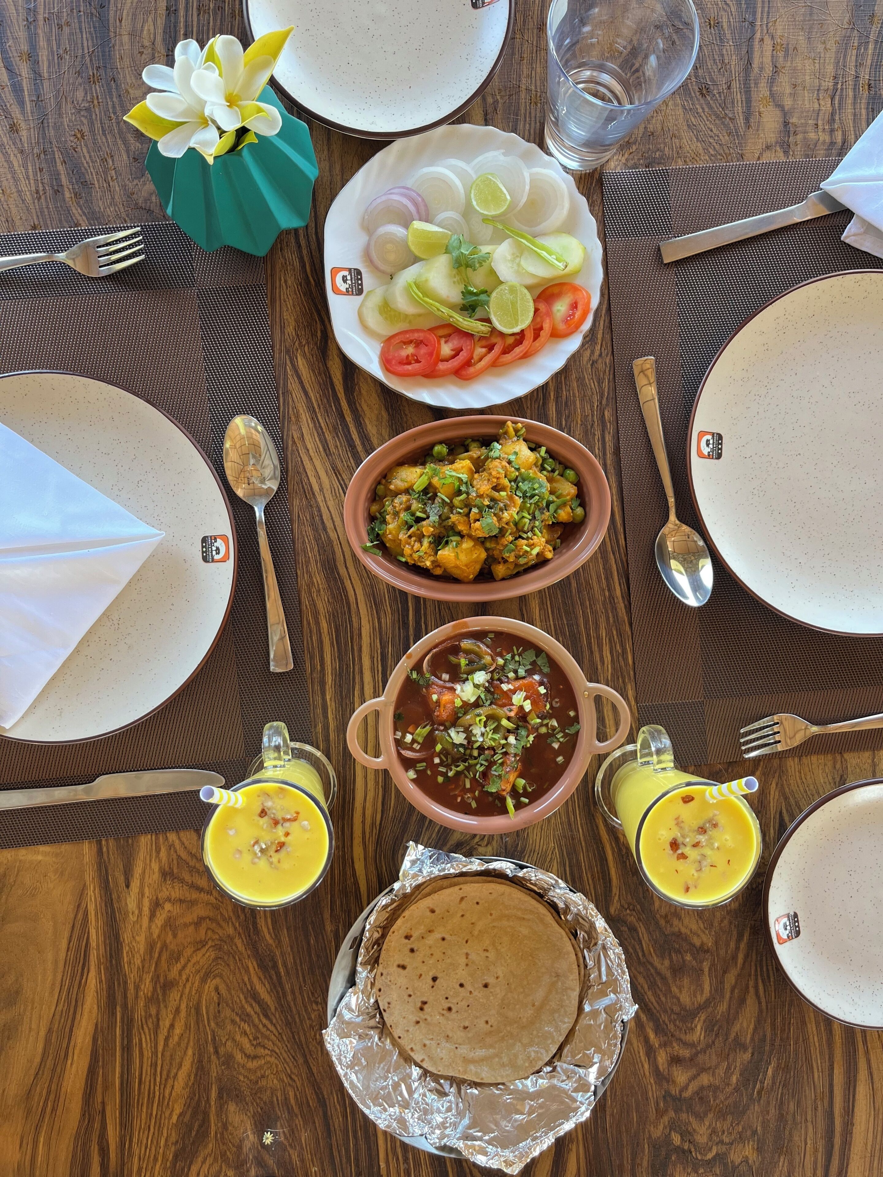 Une table de salle à manger en bois, mise avec des assiettes, des couverts, un bol de nourriture, un verre de jus de fruits et une galette plate emballée.