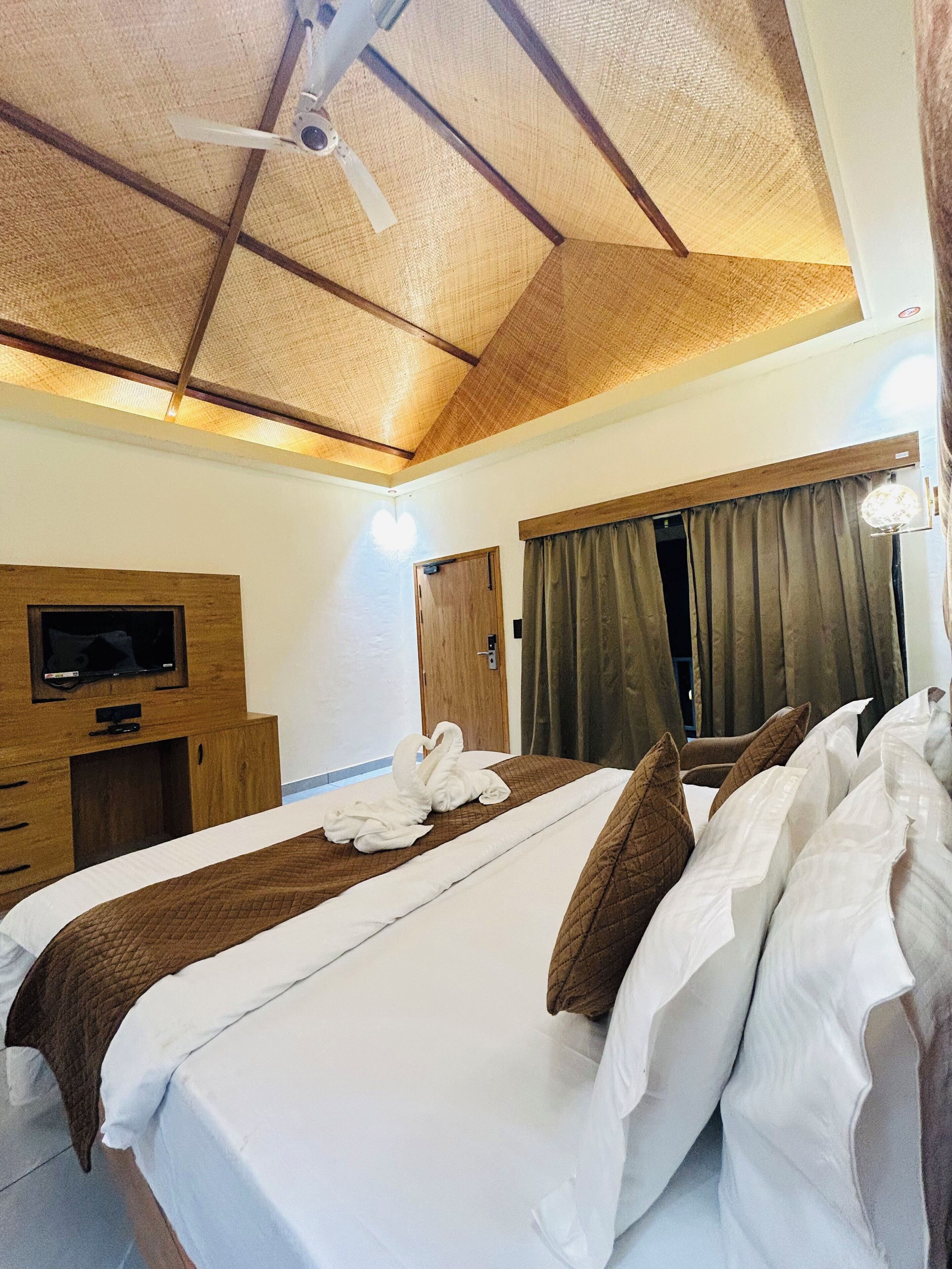 A hotel room with a bed, wooden furniture, a TV, a ceiling fan, and a window with curtains.