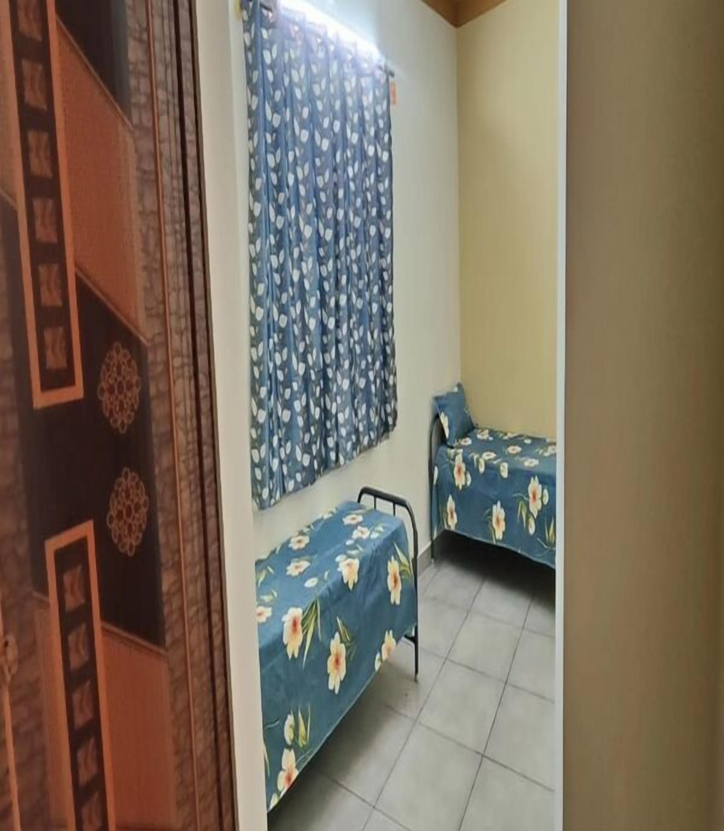 Habitación con dos camas, una a la izquierda y otra a la derecha, ambas con colchas estampadas con motivos florales. Se ve una ventana con cortina azul con diseño de hojas, y una pared con un panel decorativo a la izquierda.