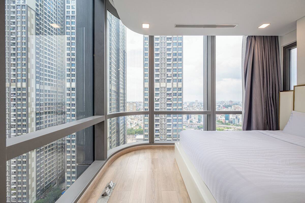 Une chambre d’hôtel moderne dotée d’un balcon incurvé, de grandes fenêtres, d’un lit et offrant une vue sur des immeubles de grande hauteur.