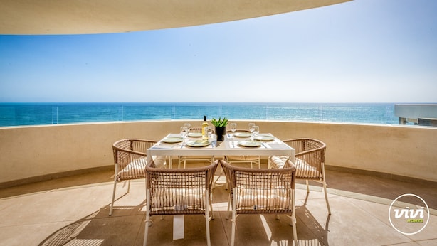 A dining table set for four with wicker chairs, a potted plant, and a view of the ocean.