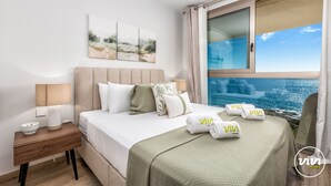 A hotel room with a bed, a nightstand, a lamp, and a view of the sea through a window.