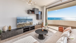 A modern living room with a large TV, a round coffee table, and a sofa. There is a balcony with a view of the ocean.