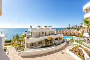 A modern building with a pool, surrounded by palm trees and a clear view of the sea.