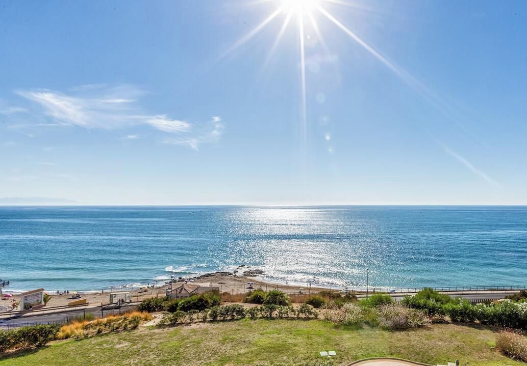 A coastal view with a clear sky, a beach, and a grassy area with a fence.