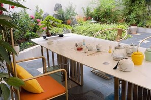 Una mesa de comedor con sillas, un juego de té y una planta en maceta en una habitación con piso de baldosas y vista a follaje exterior.