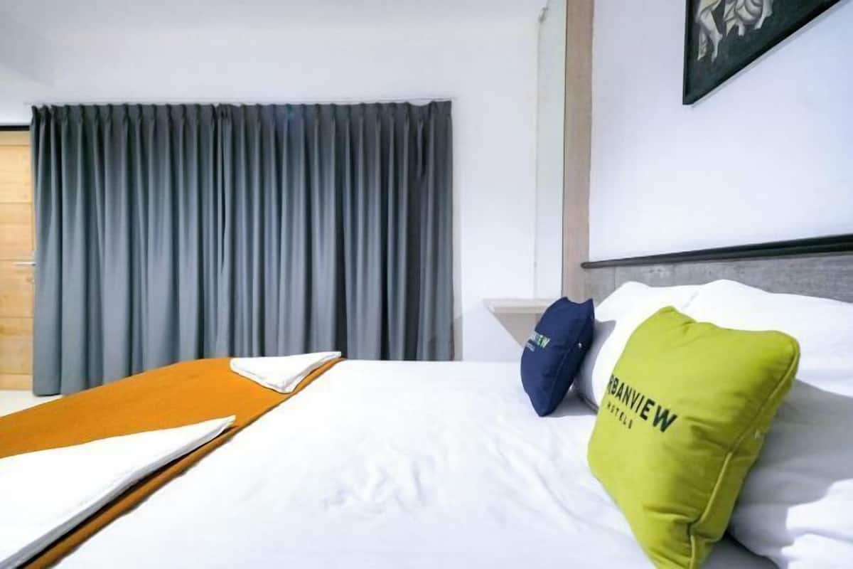 Une chambre d’hôtel avec un lit, une fenêtre avec des rideaux, un oreiller orné d’un motif et un tableau accroché au mur.
