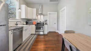 Une cuisine de taille réduite avec des meubles blancs, des appareils en acier inoxydable, une table à manger en bois et un sol à carreaux.
