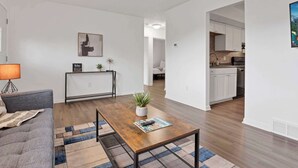 Een moderne woonkamer met een bank, salontafel en een kleine bijzettafel met een lamp. Achtergrond toont een keuken.