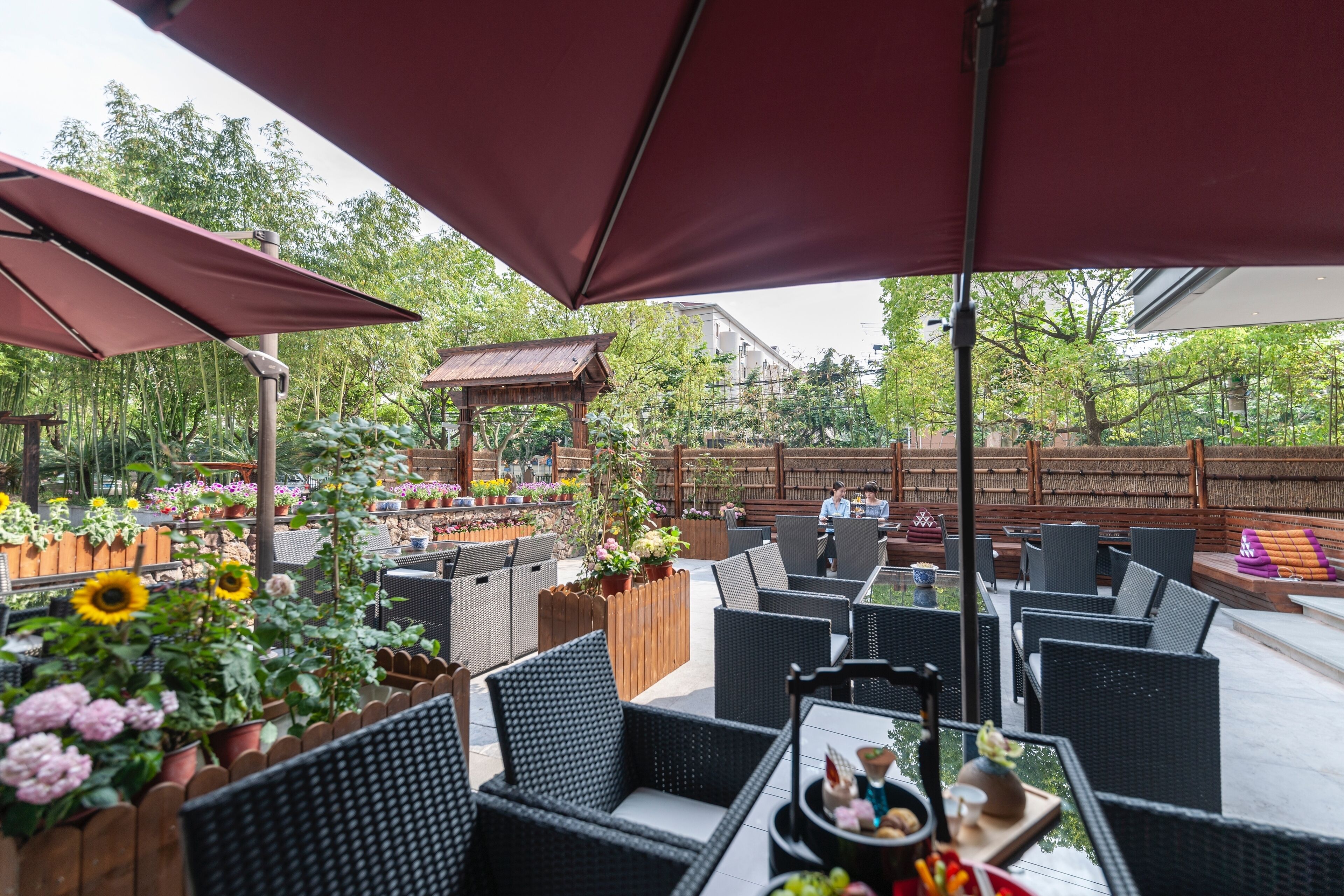 Een terras buiten met rieten meubilair, rode parasols en plantenpotten.
