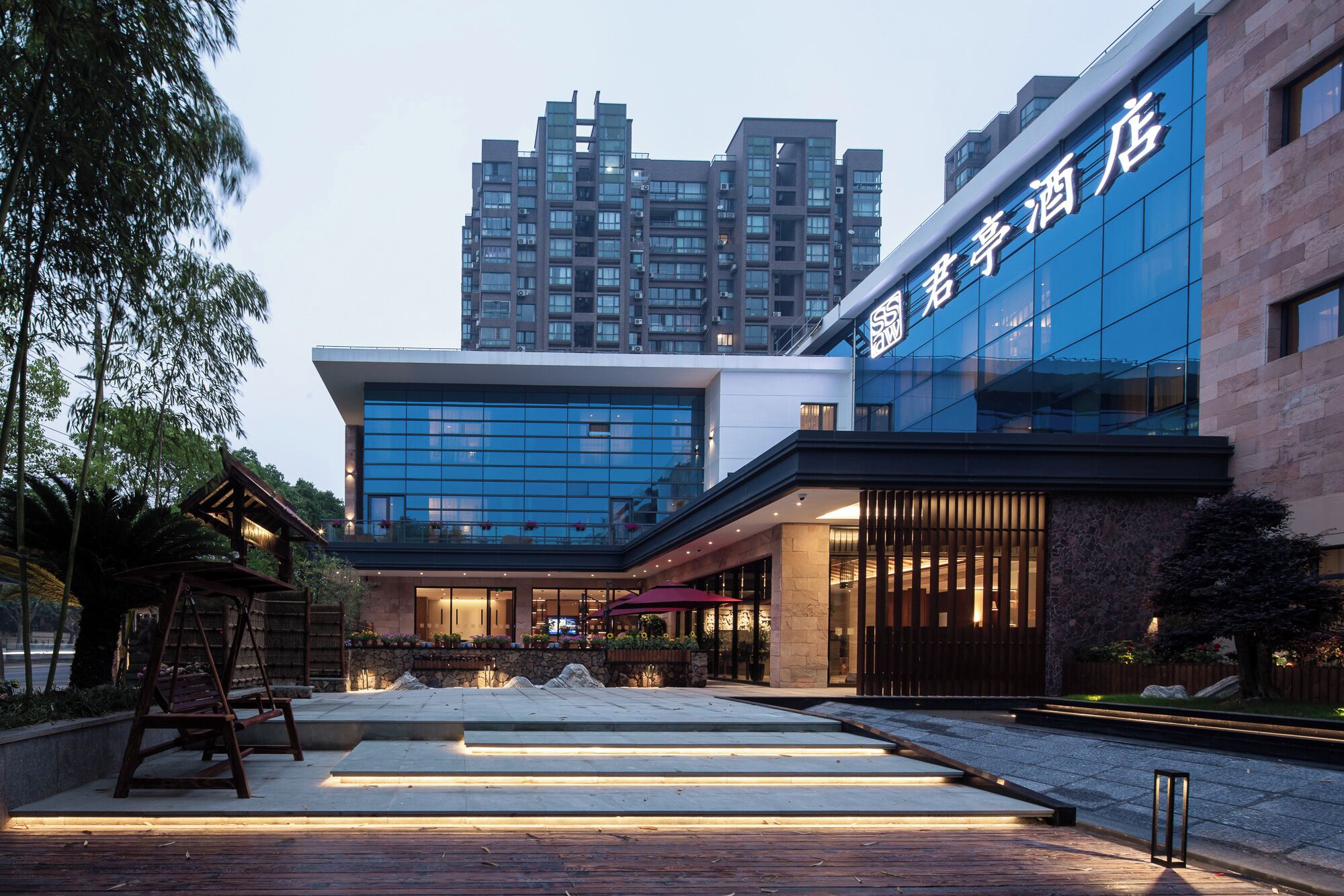 Egy modern hotel, üveg homlokzattal, kültéri ülőhellyel és egy kőből készült ösvénnyel, amely a bejárathoz vezet.