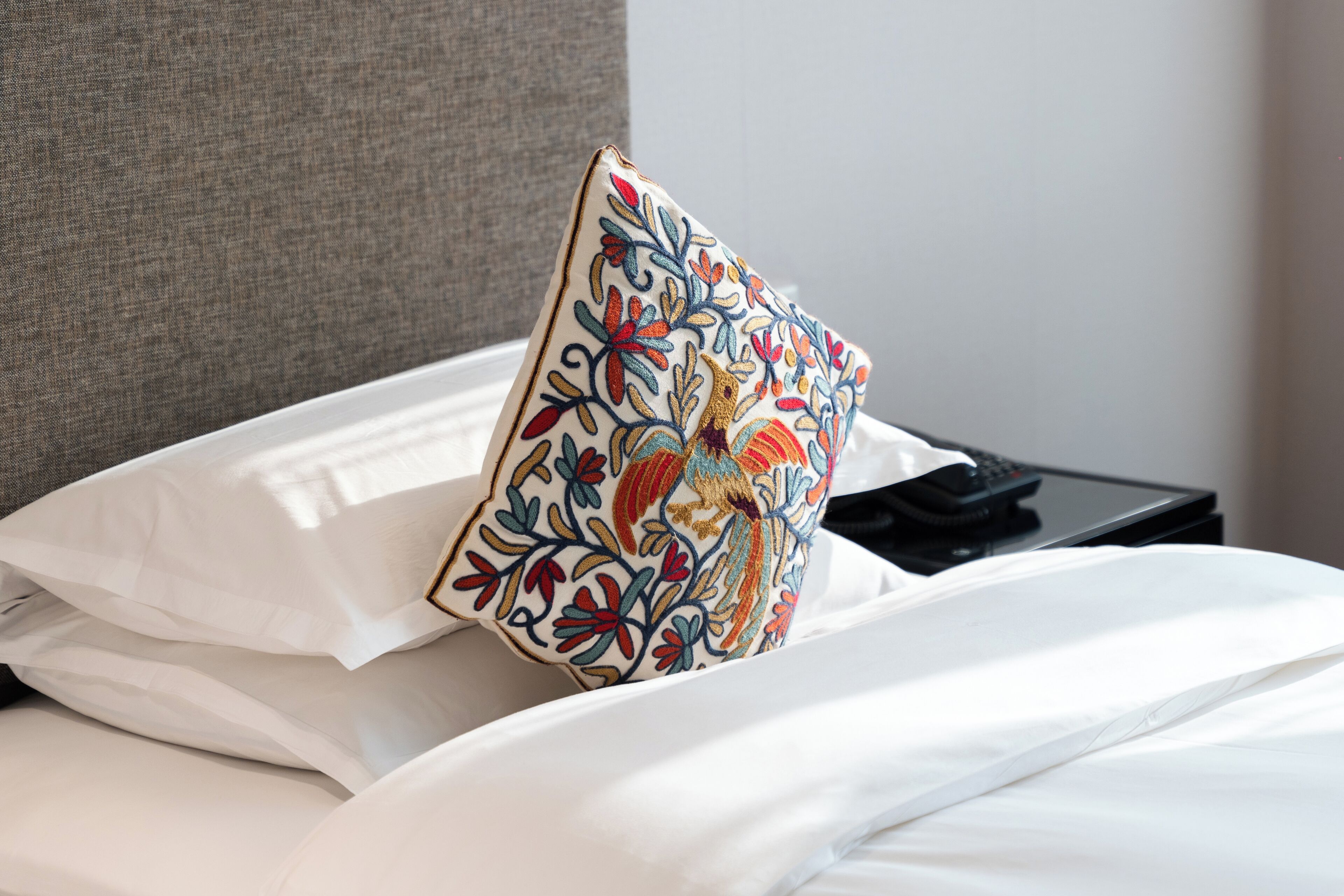 Una cama con sábanas blancas, un cabecero tapizado en gris y una almohada decorativa con bordados florales.