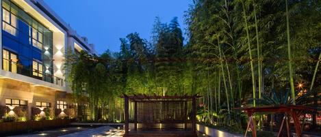 A modern building with a glass facade, a wooden pergola, and a swing set in a landscaped garden at dusk.