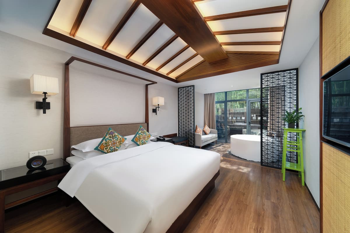 Une chambre d’hôtel moderne avec un grand lit, des tables de chevet, un bureau, une chaise, un tabouret vert, un paravent décoratif et une grande fenêtre.