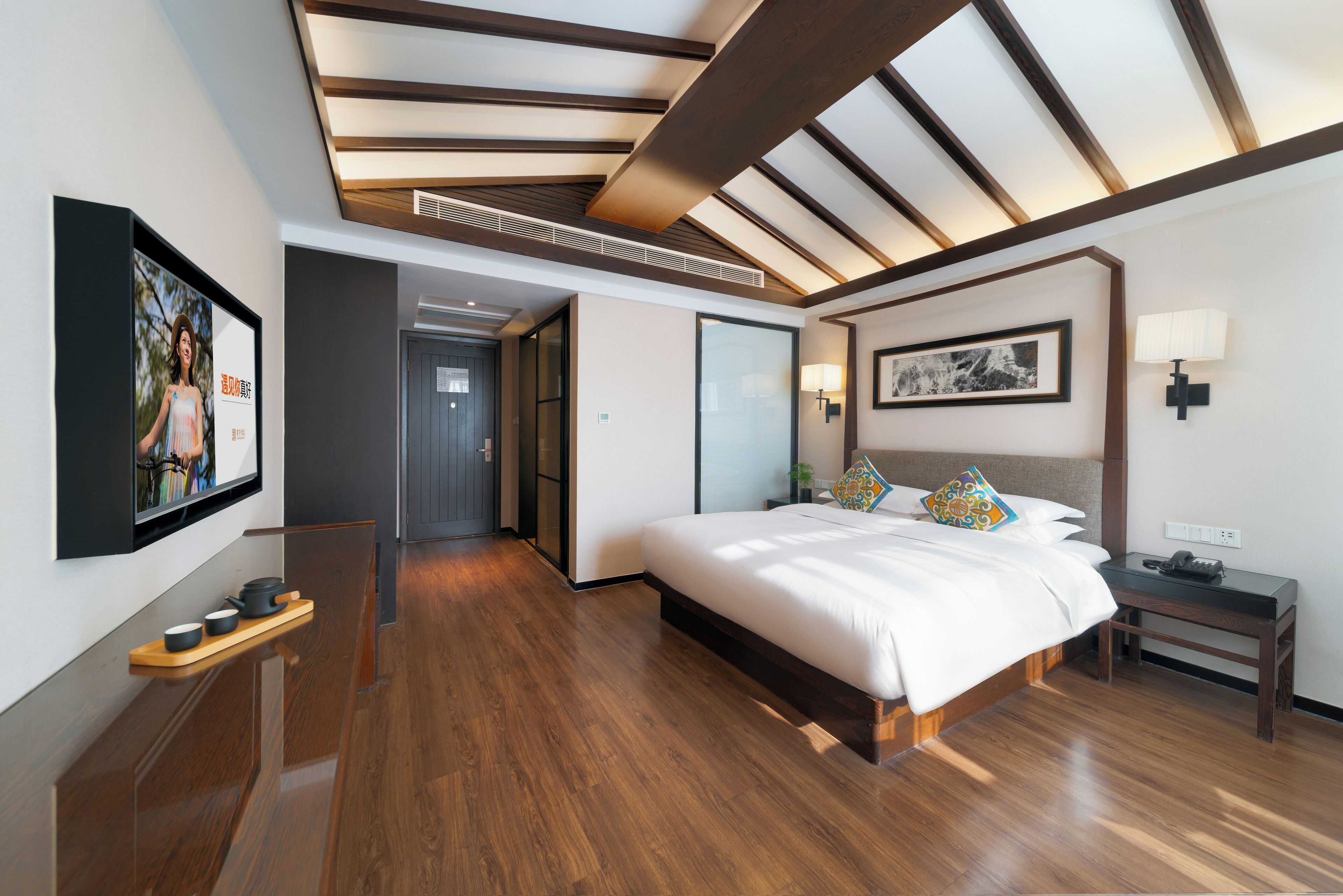 Ein modernes Hotelzimmer mit einem großen Bett, Holzboden, einer Decke mit sichtbaren Balken, einem Wand-Fernseher und einem Nachttisch mit einer Lampe.