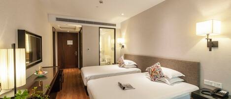 A hotel room with two beds, a TV, a bedside table with a lamp, a mirror, and a small plant.