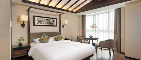 Habitación de hotel con una cama grande, lámparas de noche, una mesita con una silla y un ventanal con cortinas.