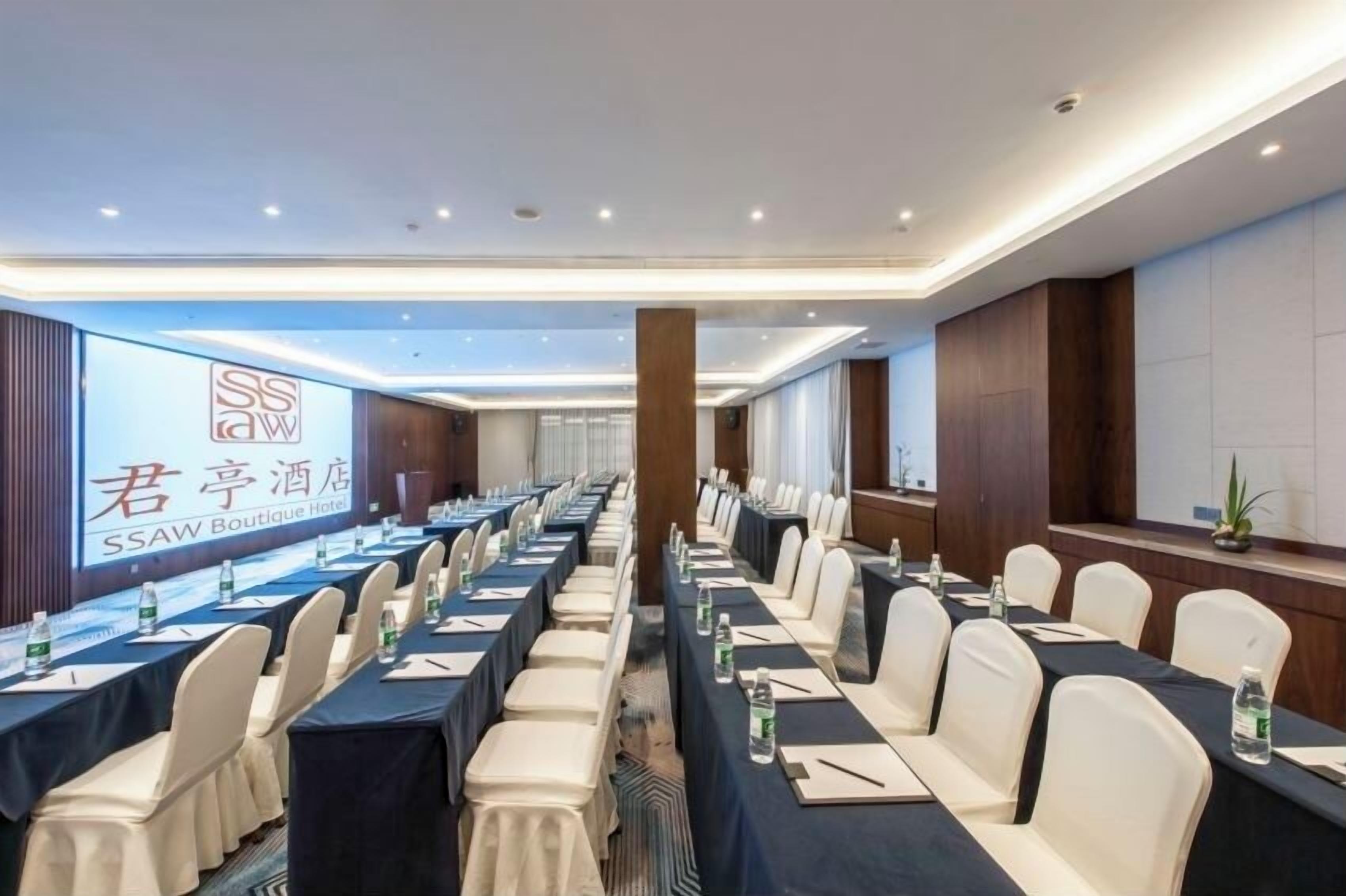 Un salón de conferencias con mesas largas, sillas y una pantalla grande que muestra el logo del Hotel SSAW Boutique.