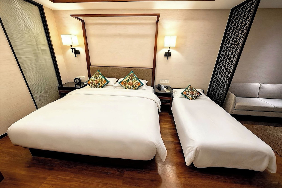 A hotel room with a large bed, a smaller bed, a bedside table with a lamp, and a sofa.