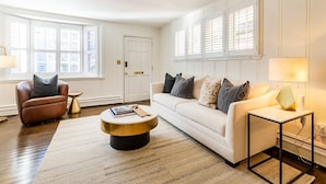 Un salon comprenant un fauteuil en cuir marron, un canapé blanc orné de coussins à motifs, une table basse ronde et une table d’appoint avec une lampe.