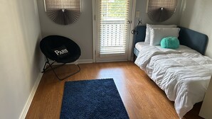 Une chambre avec un lit, un tapis bleu, une chaise sur laquelle est écrit le mot « PARIS », et deux ventilateurs fixés au mur.
