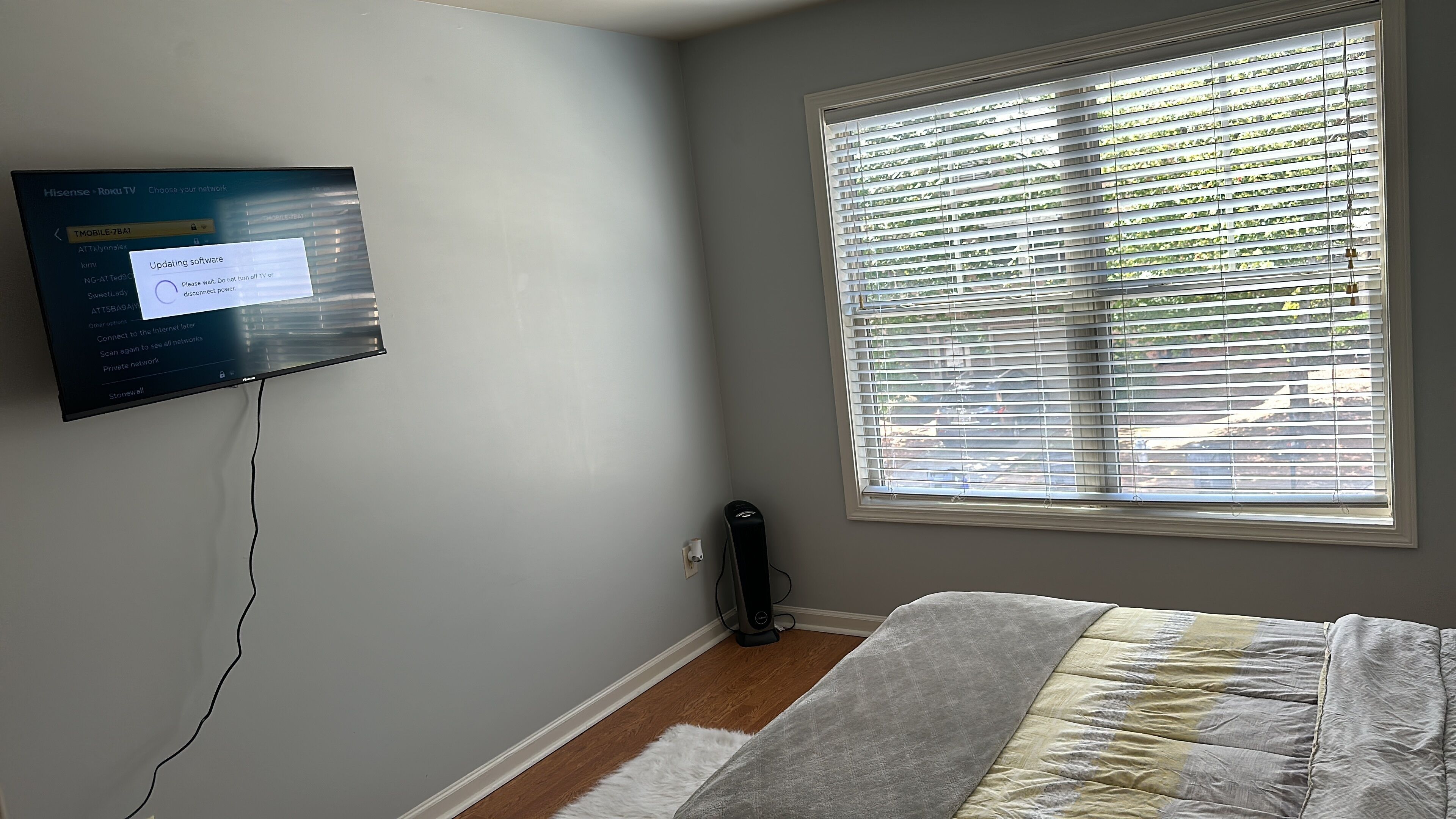 A room with a mounted television displaying a software update message, a window with blinds, a bedside table, and a bed with a blanket.