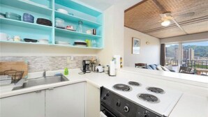 A modern kitchen with white cabinets, a stove, and a sink. There are blue shelves above the counter holding dishes and glasses. A window provides a view of the city. - Charming 2-bedroom apartment in Honolulu with AC, WiFi parking (Honolulu)