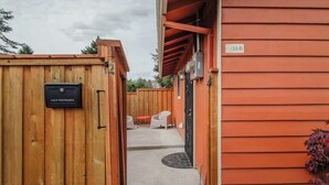 Une porte en bois avec une boîte aux lettres, un bâtiment rouge portant un numéro, et un espace patio aménagé avec du mobilier d’extérieur.