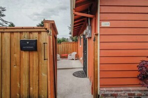 Una verja de madera con un buzón, un edificio rojo con un cartel de número, y un patio con mobiliario exterior.