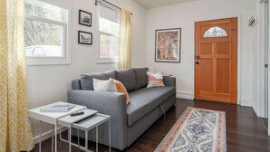 Un salon avec un canapé gris, une petite table blanche, un tapis à motifs et une porte en bois.