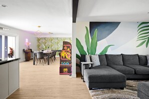 A modern living room with a grey sectional sofa, a dining area with a wooden table and chairs, and a vibrant mural with green leaves and a blue background. - The Palms Cabana | Pool, Hot Tub, Bocce & PuttPutt (Scottsdale)