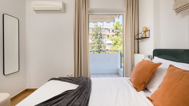 Une chambre d’hôtel avec un lit, une fenêtre avec des rideaux, un appareil de climatisation, une lampe et une vue sur les bâtiments environnants.