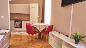 A compact living space with a kitchenette, a dining area with red chairs, a TV mounted on the wall, and a small potted plant on a white console table.