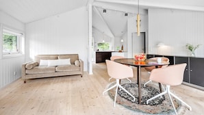 Un intérieur moderne avec un plancher en bois, un canapé beige, une table de salle à manger ronde entourée de chaises roses, et une cuisine en arrière-plan.