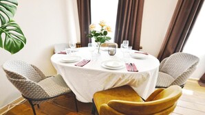 A round dining table with a white tablecloth, surrounded by four chairs, one of which is a yellow velvet chair. There is a vase with yellow flowers on the table, and the room has wooden flooring.