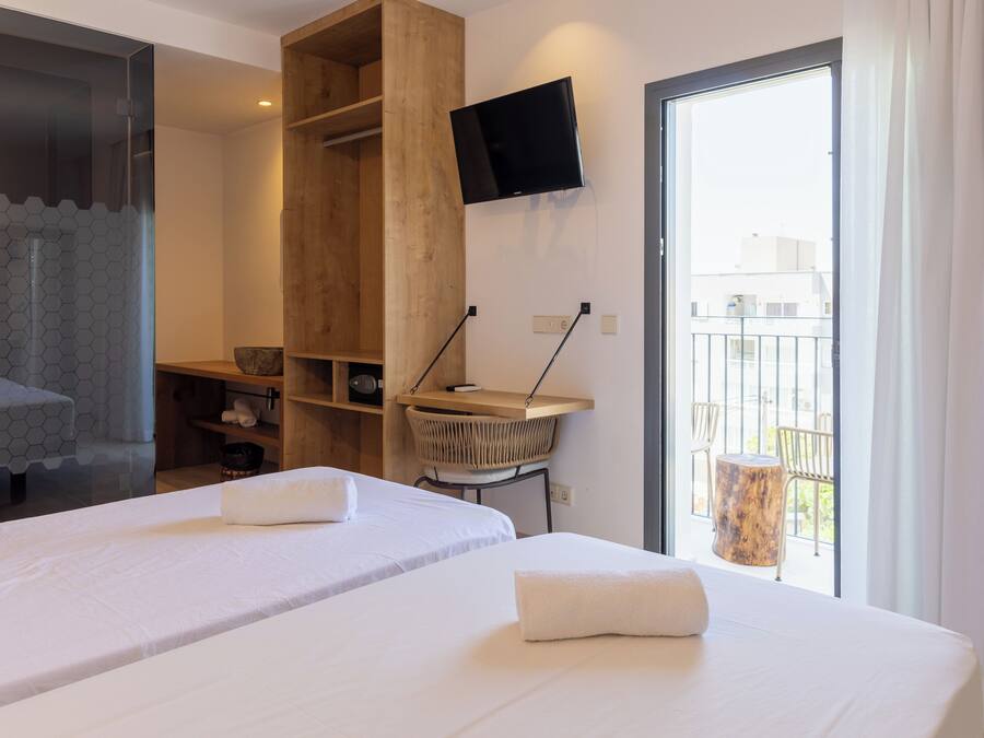 Ein Hotelzimmer mit zwei Betten, einem an der Wand befestigten Fernseher, einem Holzschrank, einem kleinen Tisch mit einem Korb und einem Balkon mit Stühlen.