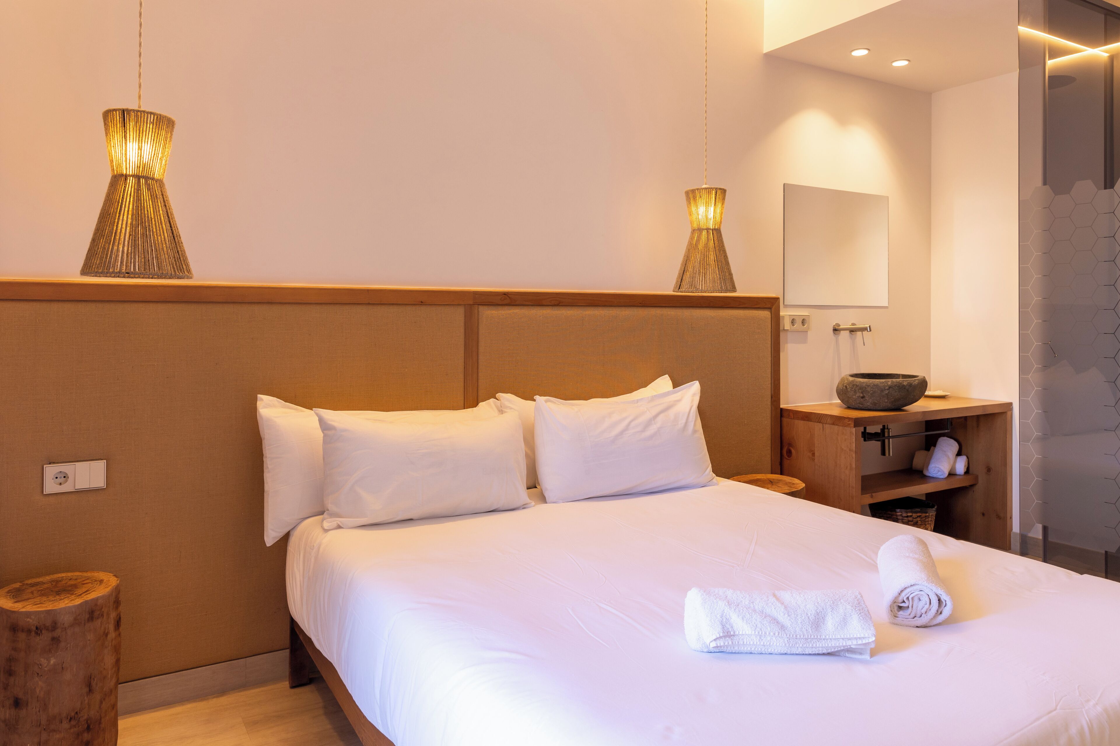 Una cama bien hecha con sábanas blancas, cabecera de madera y dos lámparas colgantes.