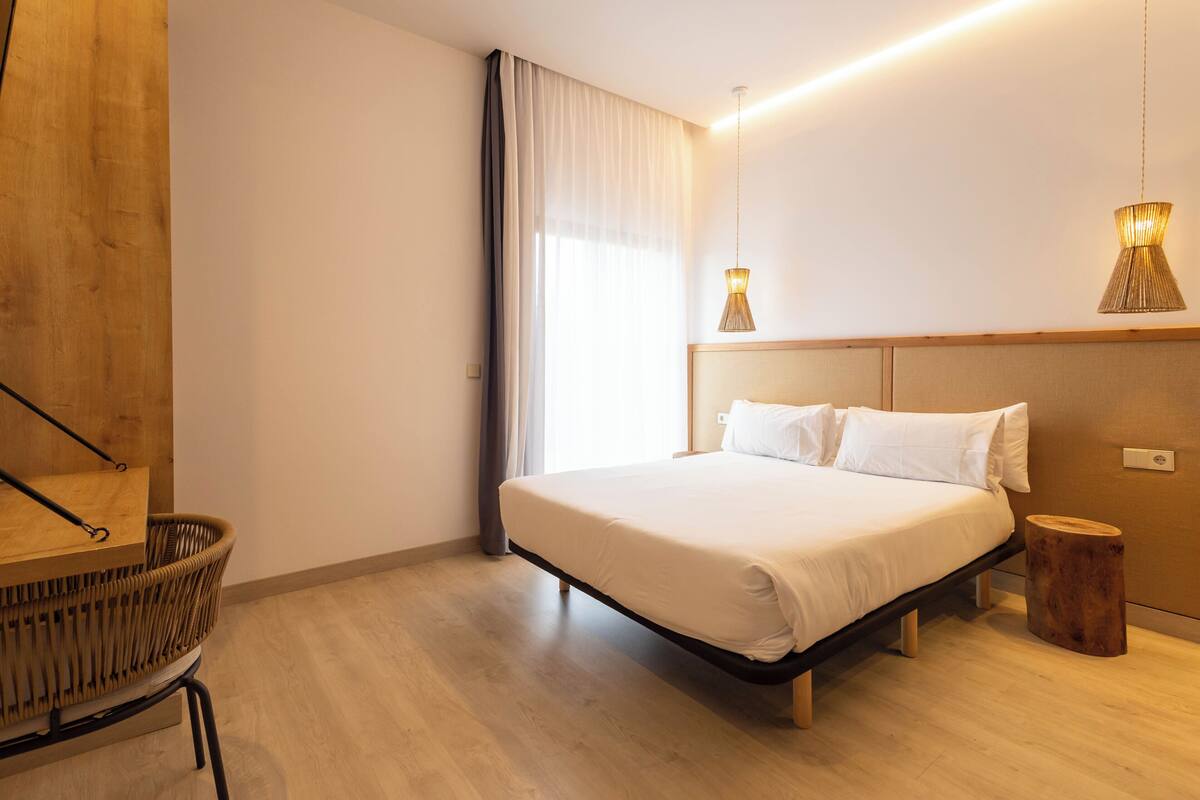 Een moderne slaapkamer met een houten hoofdbord, een groot bed, een houten nachtkastje, een rieten stoel en twee hanglampen.