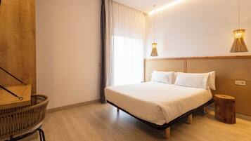 A modern bedroom with a wooden headboard, a large bed, a wooden side table, a wicker chair, and two pendant lights.