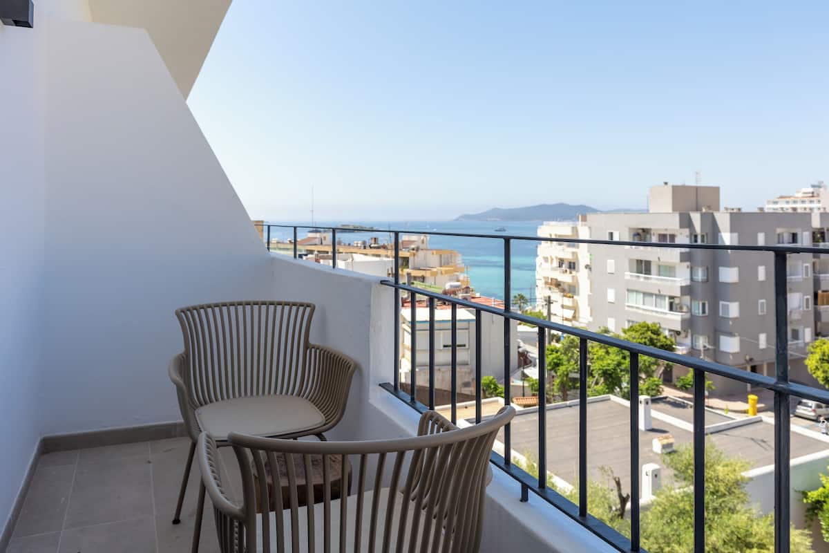 Een balkon met twee rieten stoelen, uitzicht op zee en gebouwen op de achtergrond.